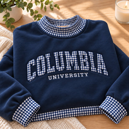 A navy blue sweatshirt with a blue and white gingham patterned collar, cuffs, and hem, featuring 'COLUMBIA UNIVERSITY' printed in white across the chest. The sweatshirt is styled on a wooden table next to a plant and a candle.