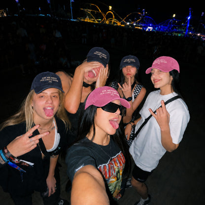 Four young women taking a selfie at night at what appears to be an outdoor music festival or event, wearing matching dark and pink caps with white text that reads 'ALL STAR FESTIVAL 2025'. The background is dark with colorful event lights in the distance.