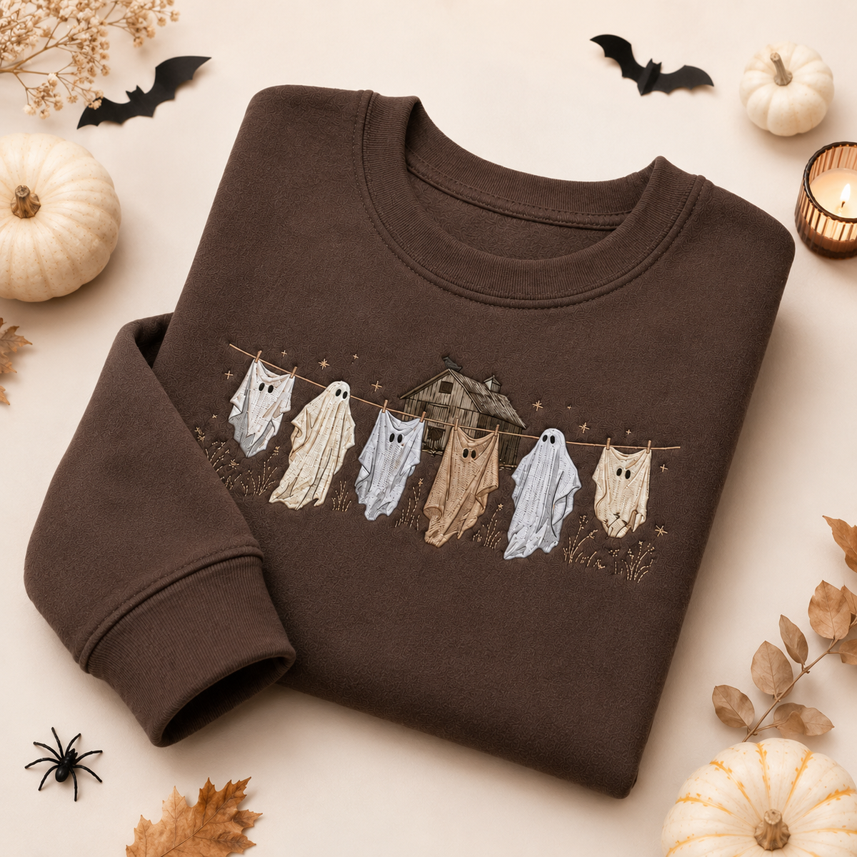 Dark brown embroidered Halloween sweatshirt featuring whimsical ghosts hanging on a clothesline with a rustic haunted house design, styled on a flat lay with autumn pumpkins, bats, and dried leaves.