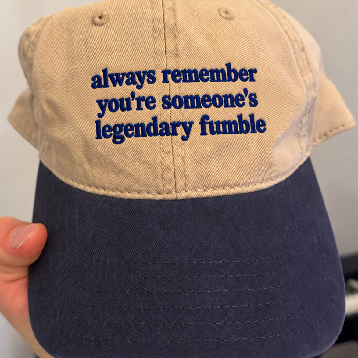 A close-up of a baseball cap being held by a hand. The cap has a beige crown with navy blue embroidered text reading 'always remember you're someone's legendary fumble' and a matching navy blue brim.