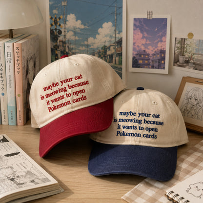 Two embroidered baseball caps displayed on a tabletop in a styled room setting. The cap in front is cream with a red brim and red text reading “maybe your cat is meowing because it wants to open Pokemon cards.