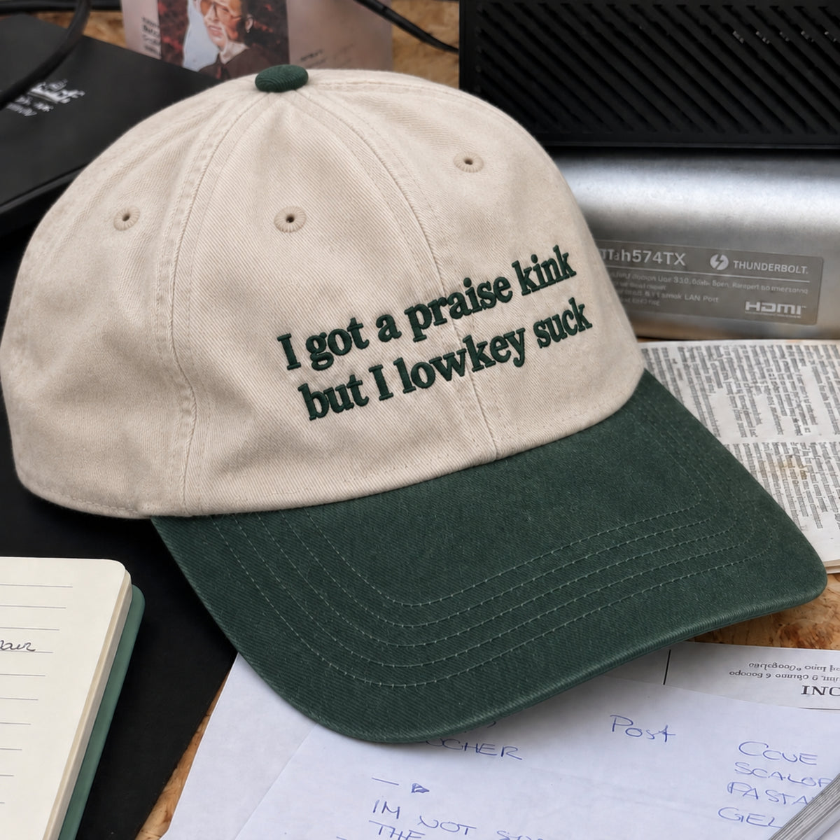 A beige and dark green baseball cap with embroidered text reading 'I got a praise kink but I lowkey suck', displayed on a desk with papers and tech accessories in the background.