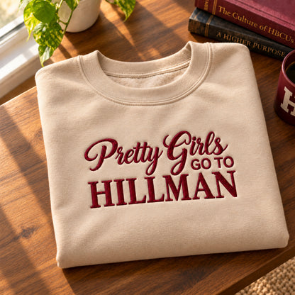 A folded cream-colored crewneck sweatshirt with 'Pretty Girls Go To HILLMAN' embroidered in dark red across the front, displayed on a wooden table with books and a plant in the background.