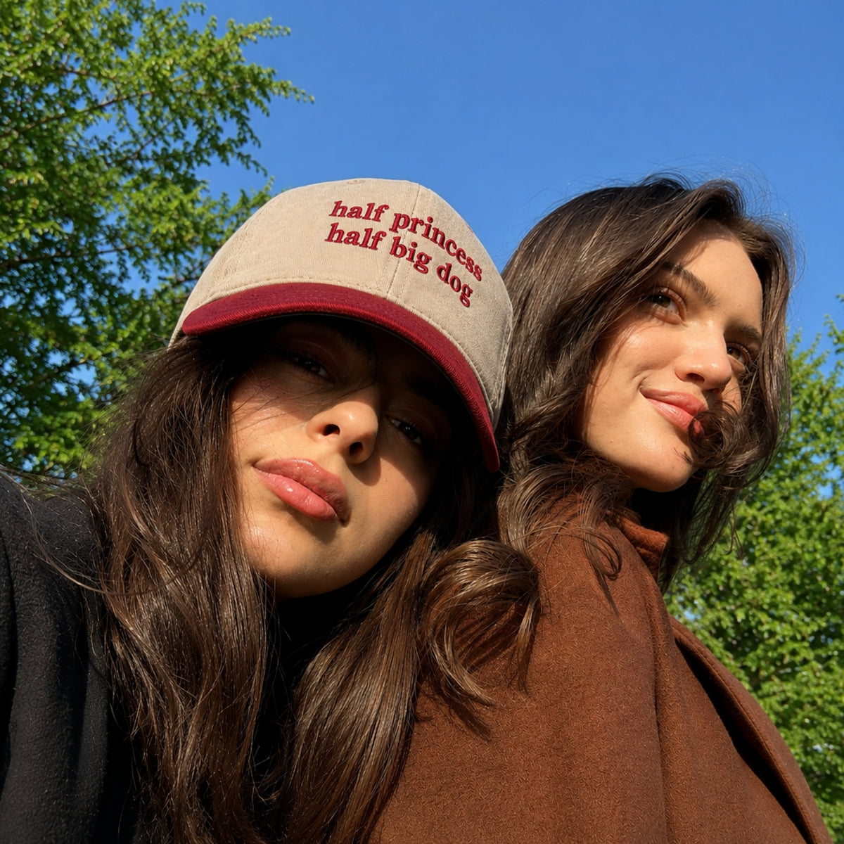 Two women outdoors under a blue sky and green trees, with one wearing a beige trucker hat with red embroidered text that reads 'half princess half big dog'.