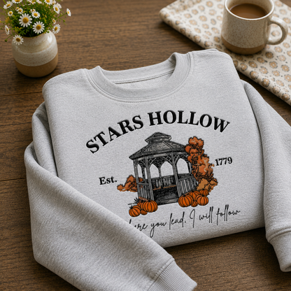 A folded light gray crewneck sweatshirt with a 'STARS HOLLOW' autumn-themed graphic print, styled on a wooden tabletop with a small potted daisy, a mug of coffee, and a patterned cloth.