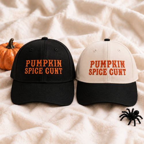Two baseball caps displayed on a cream fabric backdrop with a small orange pumpkin and black spider decoration. One cap is black with orange embroidered text reading 'PUMPKIN SPICE GUNT', and the other is off-white with black brim and black top button, with orange embroidered text reading 'PUMPKIN SPICE GUNT'.