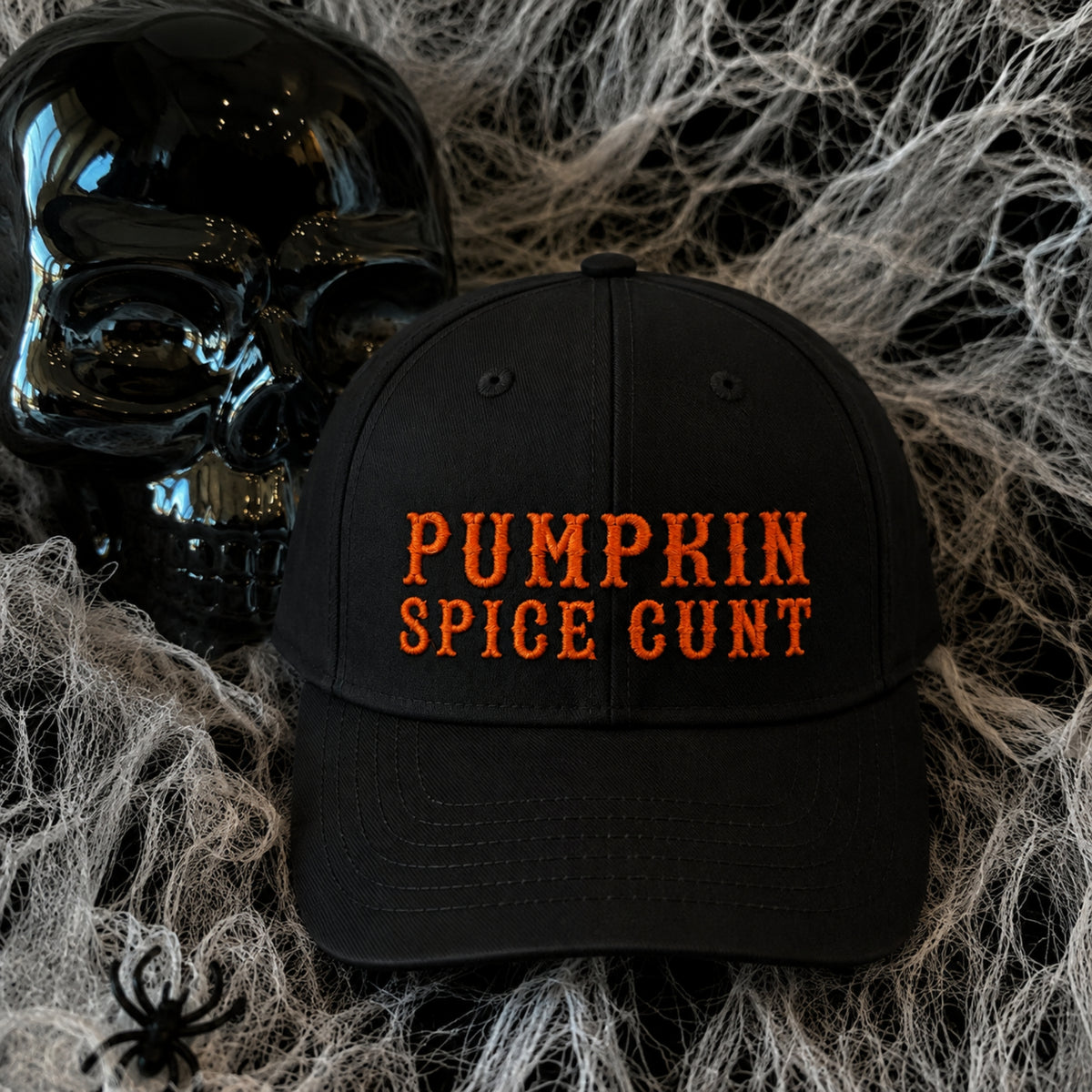 A black baseball cap with 'PUMPKIN SPICE CUNT' embroidered in orange across the front, styled on a bed of white faux spiderwebs next to a black skull.