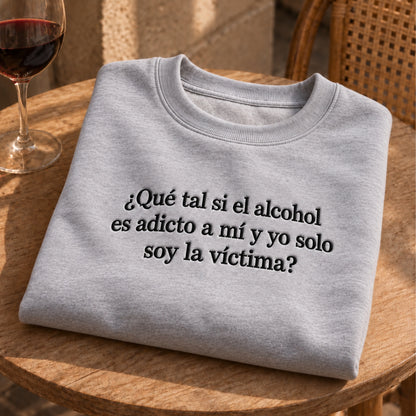 Gray embroidered sweatshirt featuring the Spanish quote “¿Qué tal si el alcohol es adicto a mí y yo solo soy la víctima?” folded on a wooden table beside a glass of red wine.