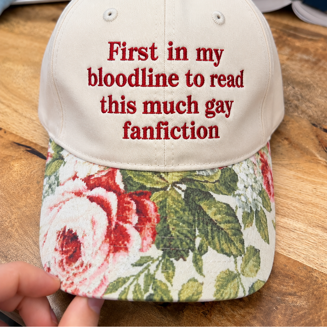 A beige baseball cap with red embroidered text reading 'First in my bloodline to read this much gay fanfiction' on the front, and a floral brim with red roses and green leaves. A hand is holding the brim, and the cap is photographed on a wooden surface.