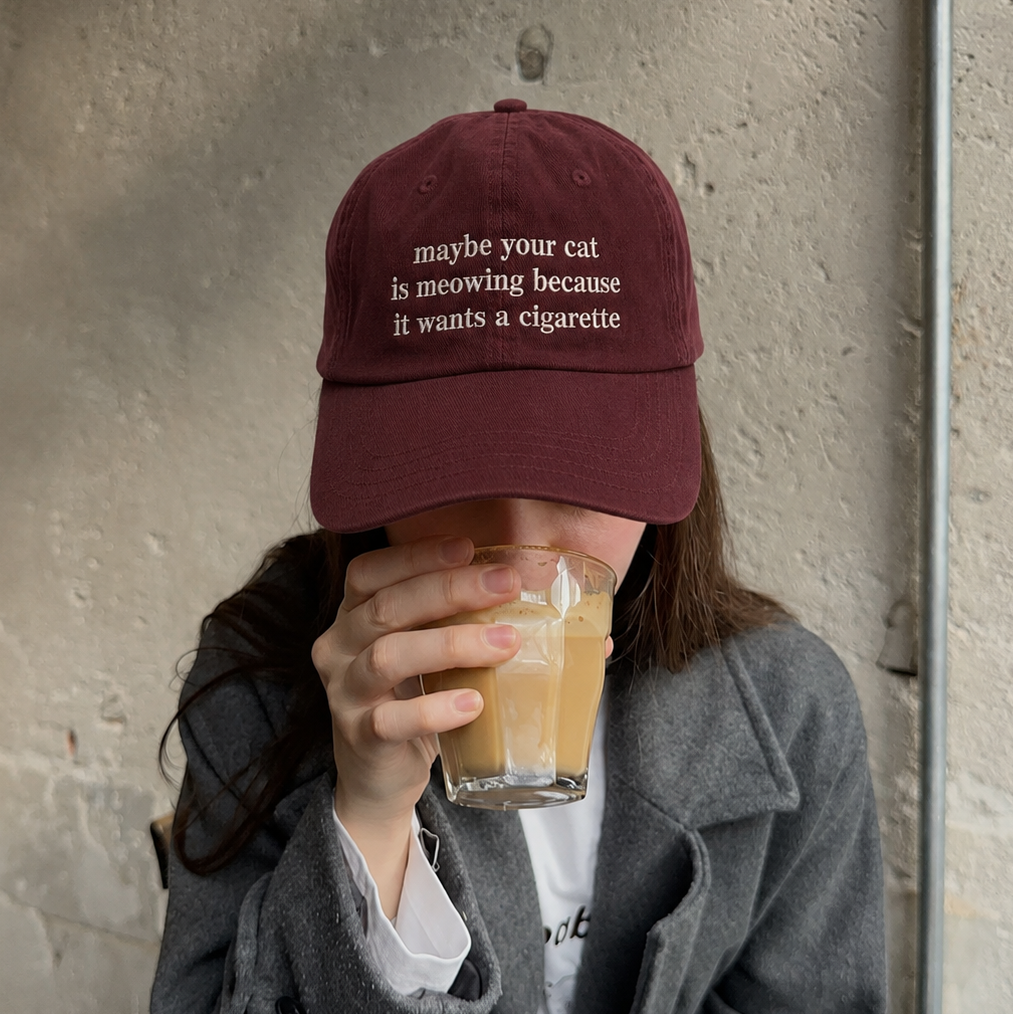 A person wearing a maroon baseball cap with white embroidered text that reads 'maybe your cat is meowing because it wants a cigarette', holding a glass of iced coffee in front of their face. They are wearing a gray coat over a white shirt, seated against a textured concrete wall.