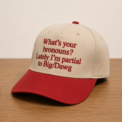 A baseball cap with a beige crown and a red brim, featuring red embroidered text that reads 'What's your bronouns? Lately I'm partial to Big/Dawg', displayed on a wooden surface against a plain beige background.