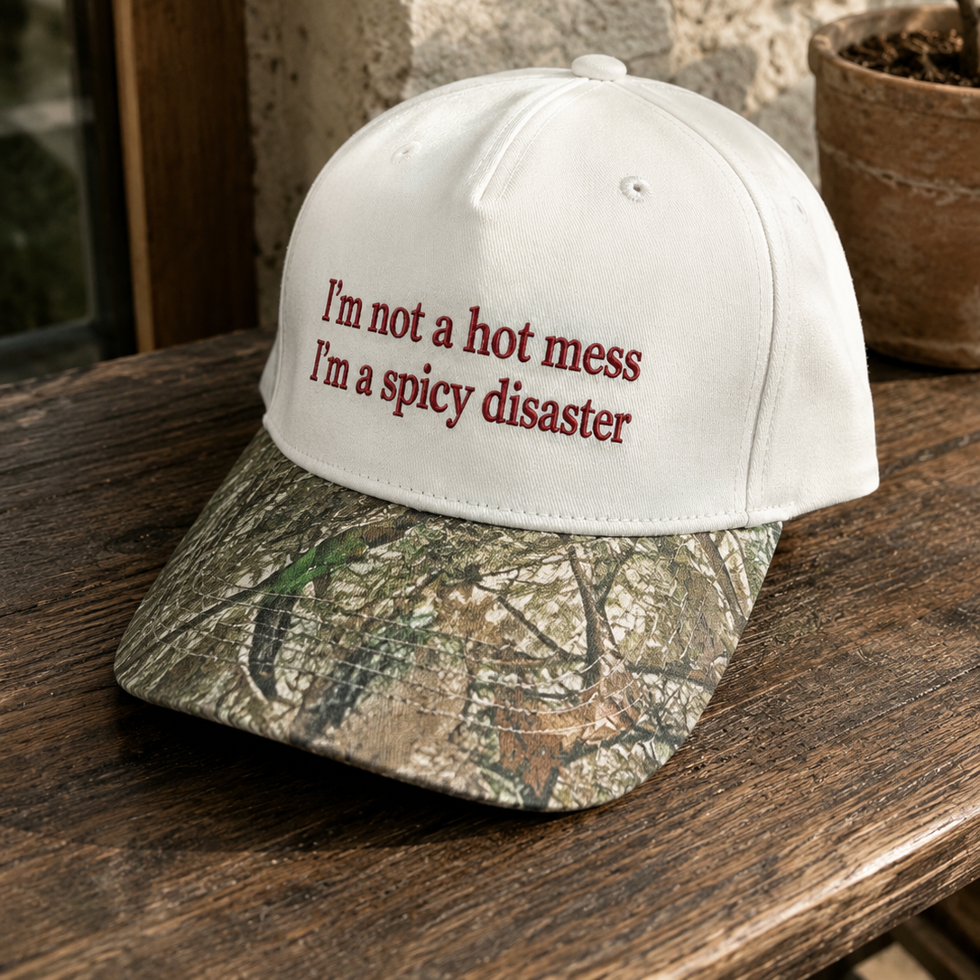 A baseball cap resting on a wooden table, featuring a white crown with the embroidered text 'I'm not a hot mess I'm a spicy disaster' in red, and a camouflage-patterned brim.