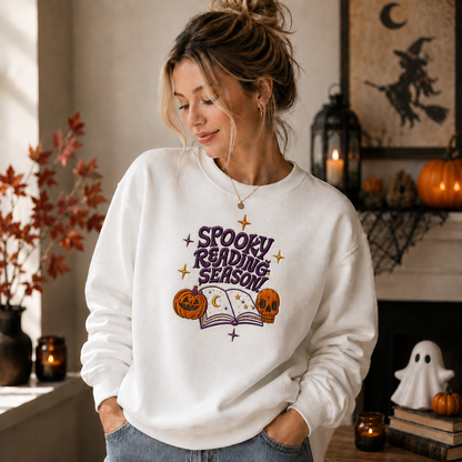 A woman wearing a white crewneck sweatshirt with a 'Spooky Reading Season' graphic featuring purple text, a purple book, and orange pumpkins, posed in a cozy, decorated Halloween-themed room.