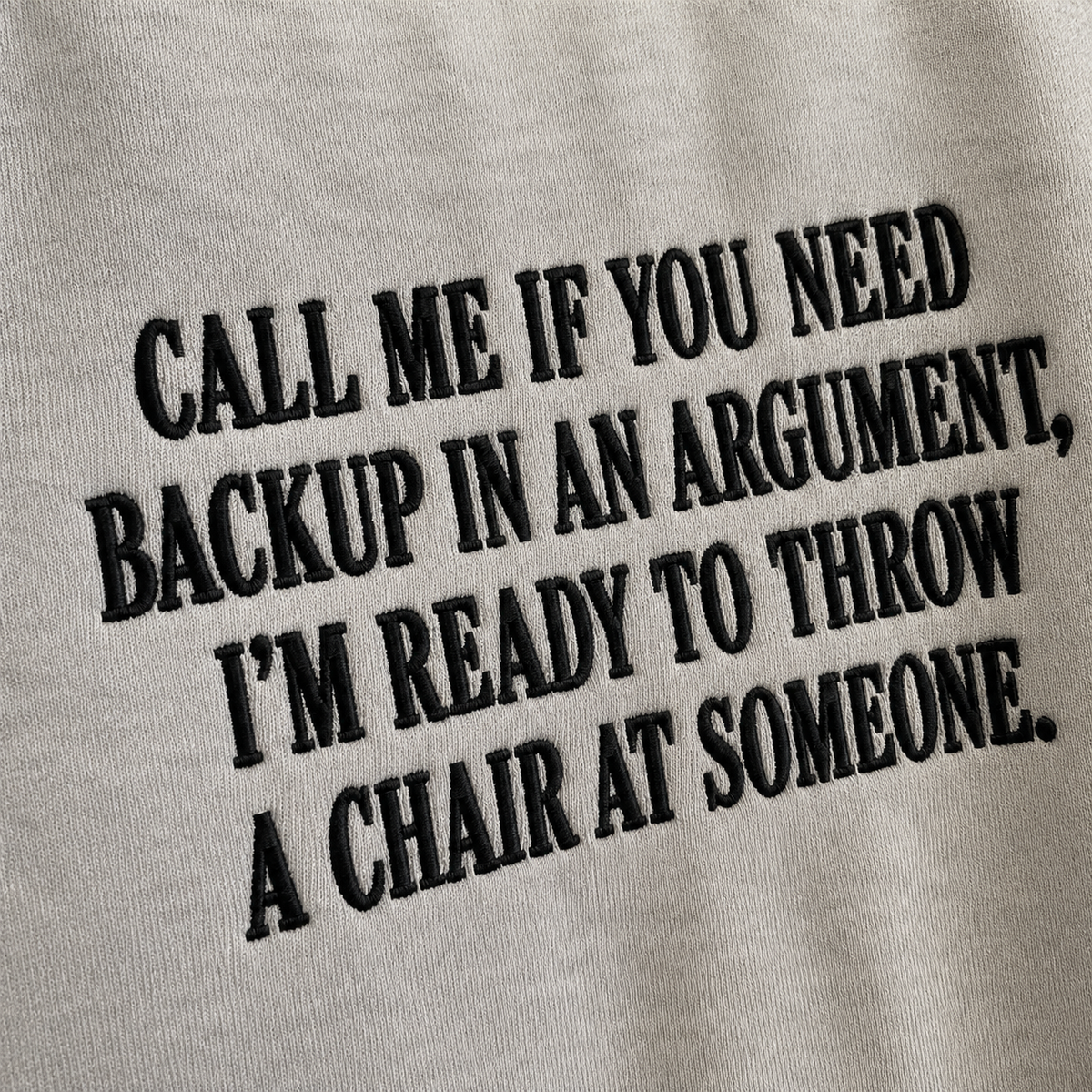 Close-up of a light gray fabric surface with bold black embroidered text reading, 'CALL ME IF YOU NEED BACKUP IN AN ARGUMENT, I'M READY TO THROW A CHAIR AT SOMEONE.'.