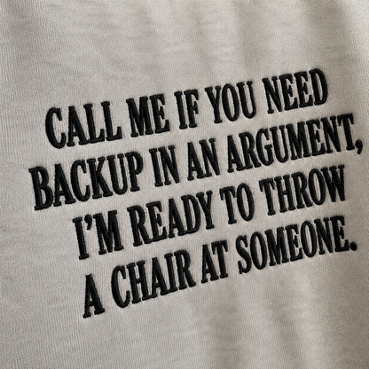 Close-up of a light gray fabric surface with bold black embroidered text reading, 'CALL ME IF YOU NEED BACKUP IN AN ARGUMENT, I'M READY TO THROW A CHAIR AT SOMEONE.'.