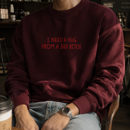 A person seated in a cafe wearing a burgundy crewneck sweatshirt with red text that reads 'I NEED A HUG FROM A BAD BITCH'.