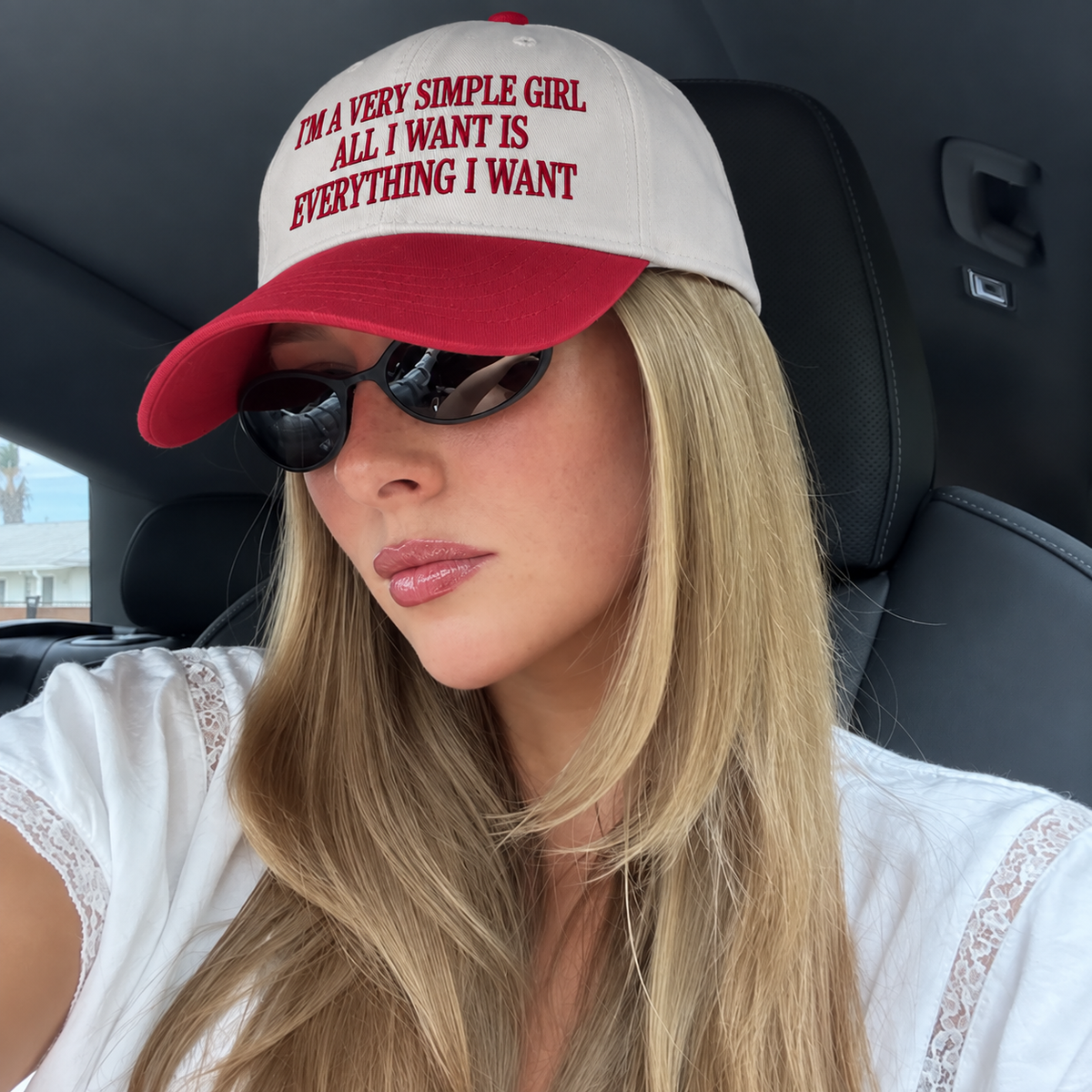 A woman wearing a red and white baseball cap with the text 'I'M A VERY SIMPLE GIRL. ALL I WANT IS EVERYTHING I WANT' in red lettering, sitting inside a car with black leather seats.