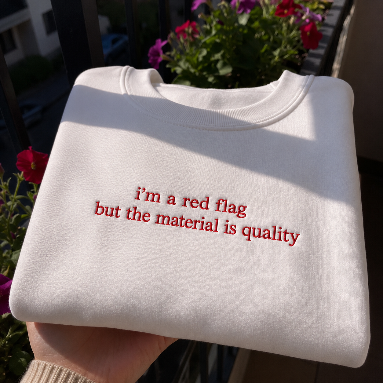 White crewneck sweatshirt held outdoors featuring red embroidered text that reads "i'm a red flag but the material is quality".
