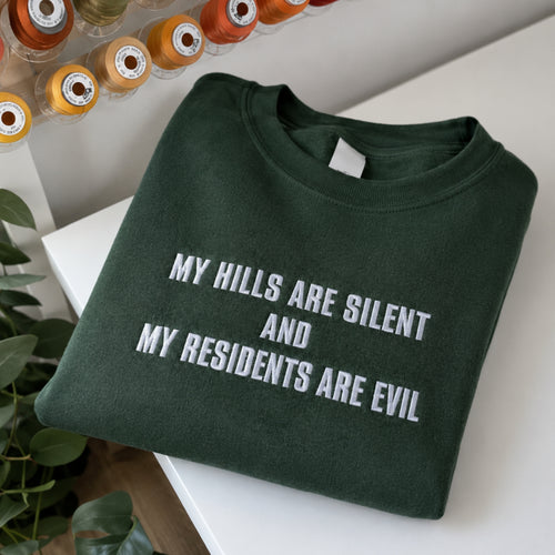 A folded dark green crewneck sweatshirt with white embroidered text reading 'MY HILLS ARE SILENT AND MY RESIDENTS ARE EVIL', displayed on a white surface with decorative thread spools and greenery in the background.