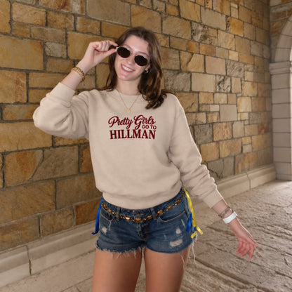 A woman wearing a beige crewneck sweatshirt with 'Pretty Girls I Go To HILLMAN' printed in maroon across the chest, paired with denim shorts, standing in front of a stone wall.