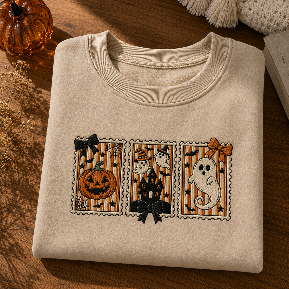 A folded cream-colored crewneck sweatshirt with a Halloween-themed graphic print, displayed on a wooden surface with autumn props.