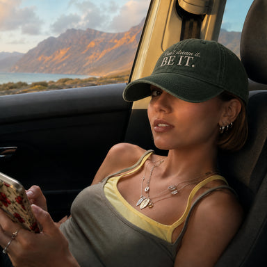 A woman sitting in a car wearing a dark green baseball cap with the embroidered text 'I can dream it. BE IT.
