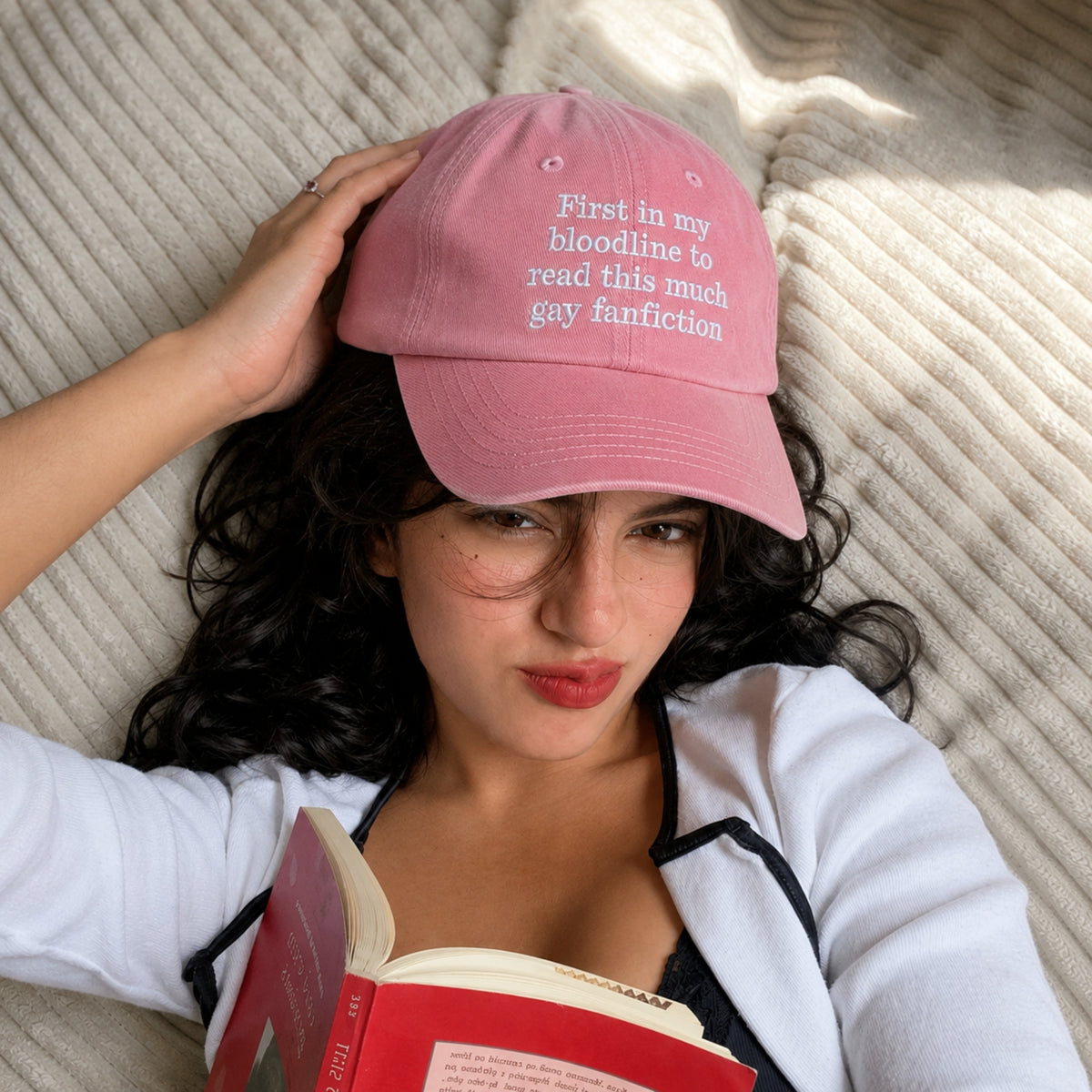 Young woman lying on a cream blanket while reading a book and wearing a pink embroidered dad hat that says "First in my bloodline to read this much gay fanfiction." The aesthetic book lover cap features white embroidered text and is styled as a funny BookTok-inspired accessory for romance readers and fanfiction fans.