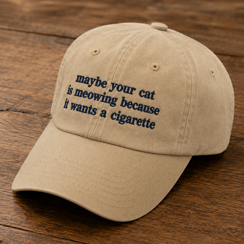 A beige baseball cap with navy blue embroidered text reading 'maybe your cat is meowing because it wants a cigarette', displayed on a dark wood background.