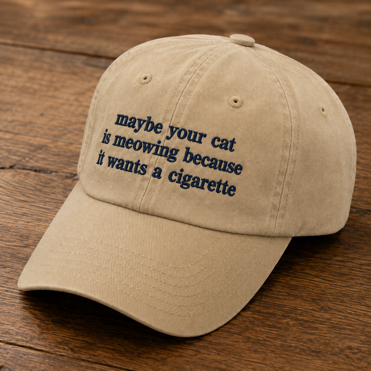A beige baseball cap with navy blue embroidered text reading 'maybe your cat is meowing because it wants a cigarette', displayed on a dark wood background.