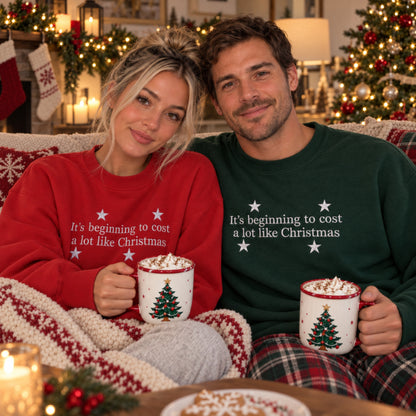 A couple sitting together on a couch wrapped in a knit blanket, each holding a mug with a Christmas tree design. The woman wears a red sweatshirt with white text reading 'It's beginning to cost a lot like Christmas,' and the man wears a dark green sweatshirt with the same text.