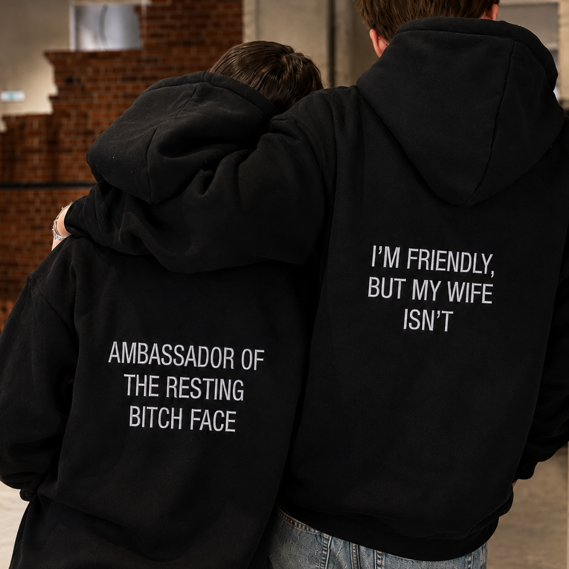 Two people standing close together indoors, shown from the back wearing matching black hoodies with white text. The hoodie on the right reads "I'M FRIENDLY, BUT MY WIFE ISN'T" and the hoodie on the left reads "AMBASSADOR OF THE RESTING BITCH FACE.