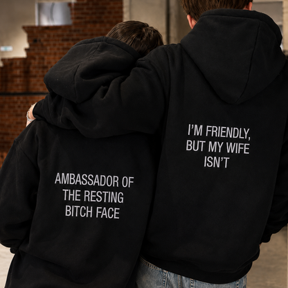 Two people standing close together indoors, shown from the back wearing matching black hoodies with white text. The hoodie on the right reads "I'M FRIENDLY, BUT MY WIFE ISN'T" and the hoodie on the left reads "AMBASSADOR OF THE RESTING BITCH FACE.