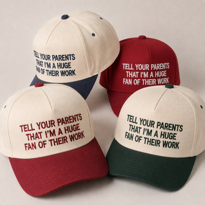 Four baseball caps arranged on a light neutral background, each with a cream crown and contrasting brim in navy, burgundy, red, and dark green. The front panels are printed with the phrase 'TELL YOUR PARENTS THAT I'M A HUGE FAN OF THEIR WORK' in matching colors.