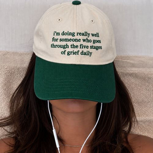 A person wearing a baseball cap with a cream crown and dark green brim. The cap has green embroidered text that reads, 'i'm doing really well for someone who goes through the five stages of grief daily.