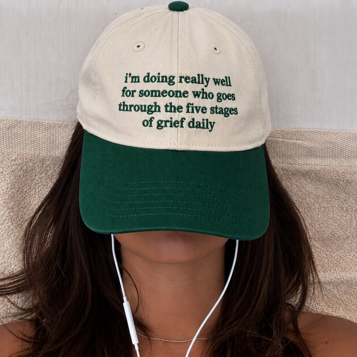 A person wearing a baseball cap with a cream crown and dark green brim. The cap has green embroidered text that reads, 'i'm doing really well for someone who goes through the five stages of grief daily.