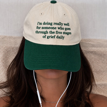 A person wearing a baseball cap with a cream crown and dark green brim. The cap has green embroidered text that reads, 'i'm doing really well for someone who goes through the five stages of grief daily.