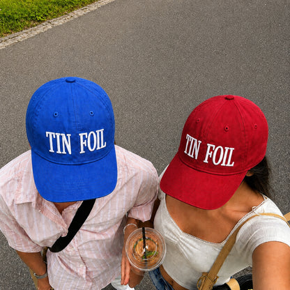 Two people outdoors wearing matching baseball caps, one blue and one red, both with 'TIN FOIL' embroidered in white across the front. The person on the left wears a light pink plaid shirt and holds a drink, while the person on the right wears a white top and a tan shoulder bag.