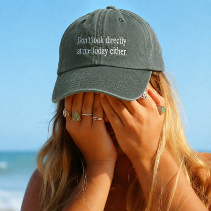 A woman on a beach wearing a dark gray baseball cap with the embroidered text 'Don't look directly at me today either' in white. She is covering her face with her hands, which are adorned with several rings, against a background of blue sky and ocean.