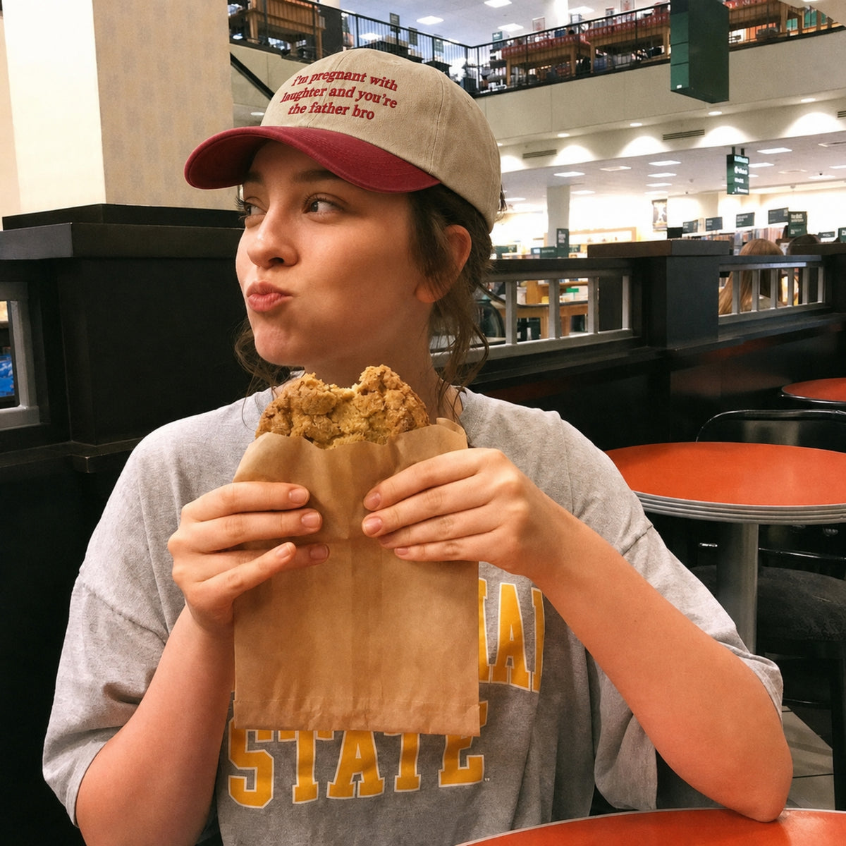 A woman in a food court setting wearing a gray and yellow 'NAA STATE' t-shirt and a beige and maroon baseball cap, eating a piece of fried chicken wrapped in a brown paper bag.