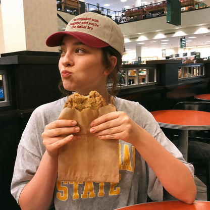 A woman in a food court setting wearing a gray and yellow 'NAA STATE' t-shirt and a beige and maroon baseball cap, eating a piece of fried chicken wrapped in a brown paper bag.