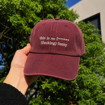 A hand holds a maroon baseball cap outdoors with green trees and a building in the background. The cap has white embroidered text reading 'this is so f****** (fucking) funny'.