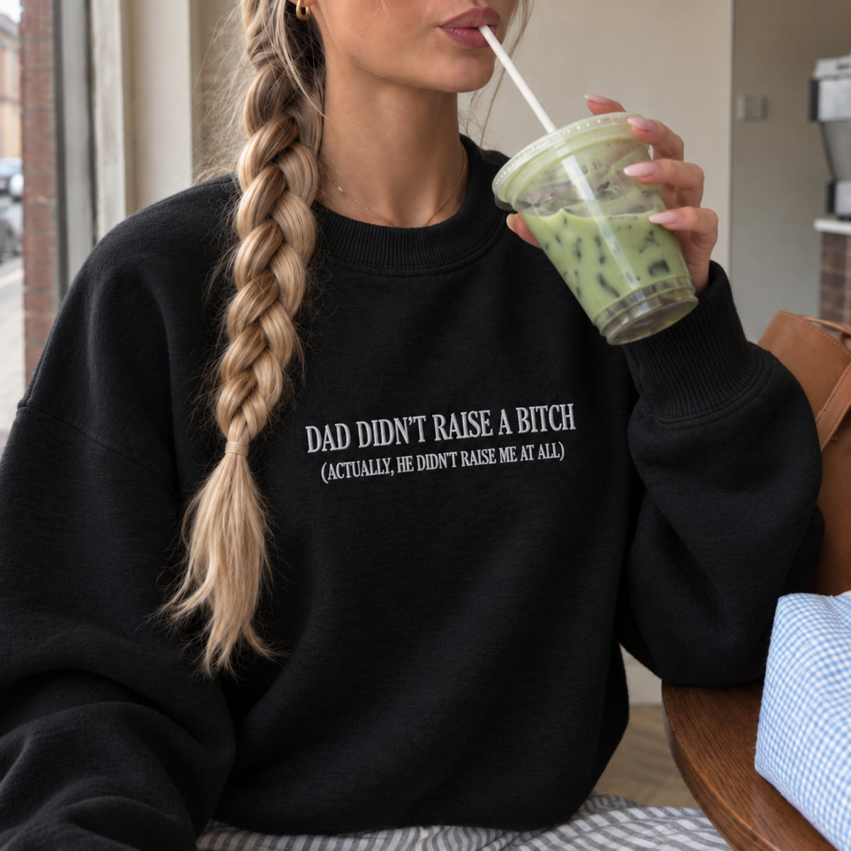 A woman wearing a black crewneck sweatshirt with white text that reads 'DAD DIDN'T RAISE A BITCH (ACTUALLY, HE DIDN'T RAISE ME AT ALL)'. She is holding a green iced drink in a cafe setting.