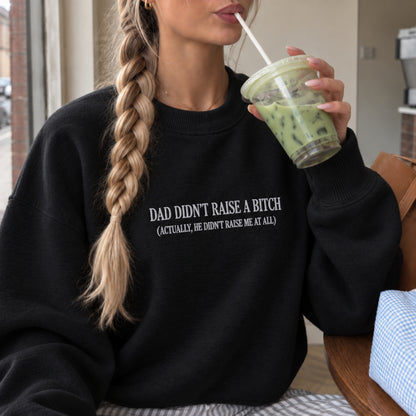 A woman wearing a black crewneck sweatshirt with white text that reads 'DAD DIDN'T RAISE A BITCH (ACTUALLY, HE DIDN'T RAISE ME AT ALL)'. She is holding a green iced drink in a cafe setting.