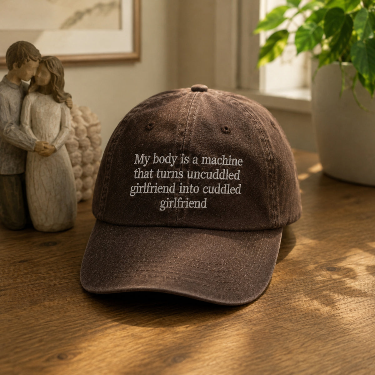 A dark brown distressed baseball cap with white embroidered text reading 'My body is a machine that turns uncuddled girlfriend into cuddled girlfriend' resting on a wooden table in a home setting.