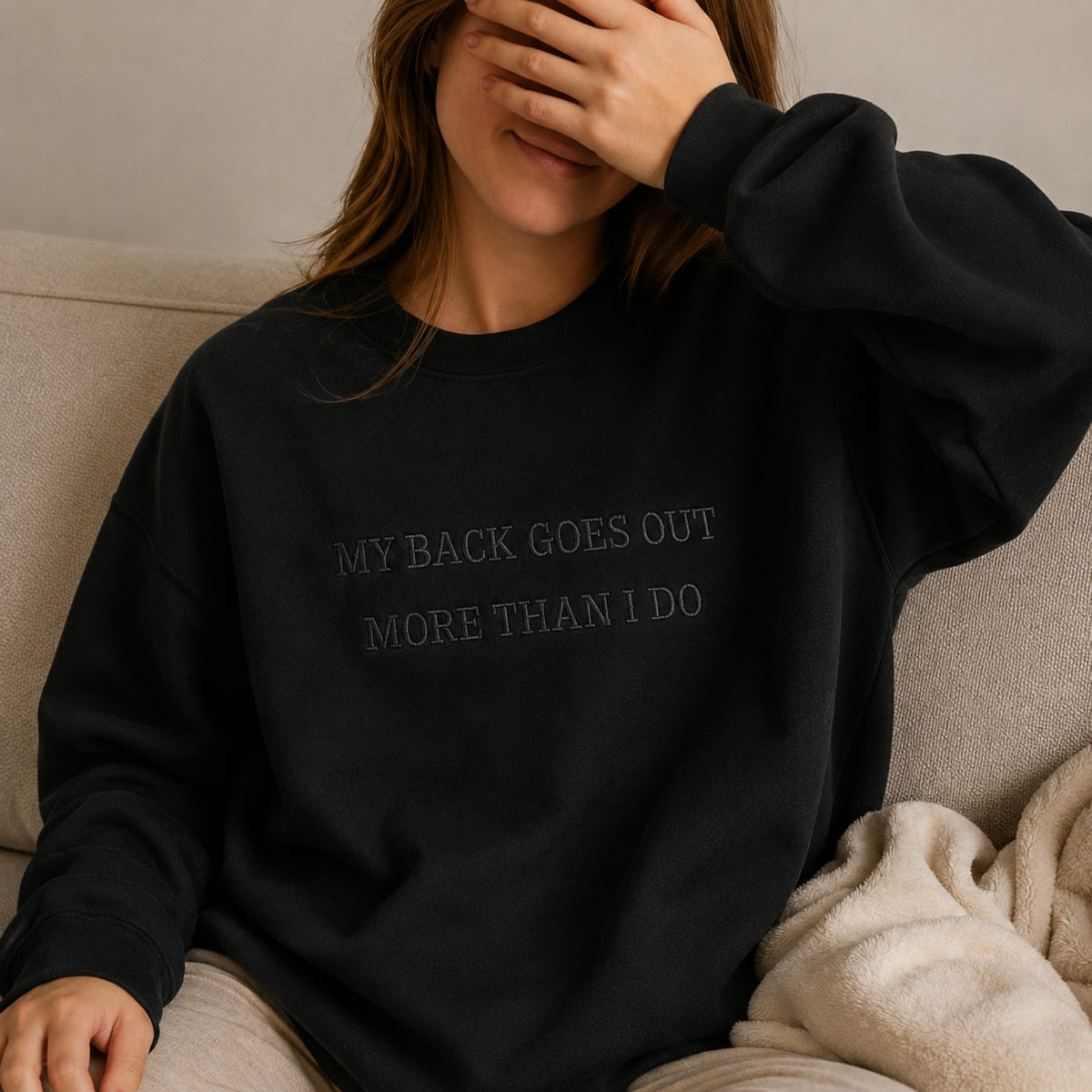 A woman wearing a black crewneck sweatshirt with the text 'MY BACK GOES OUT MORE THAN I DO' in a dark gray tone, sitting on a beige couch with a cream throw blanket.