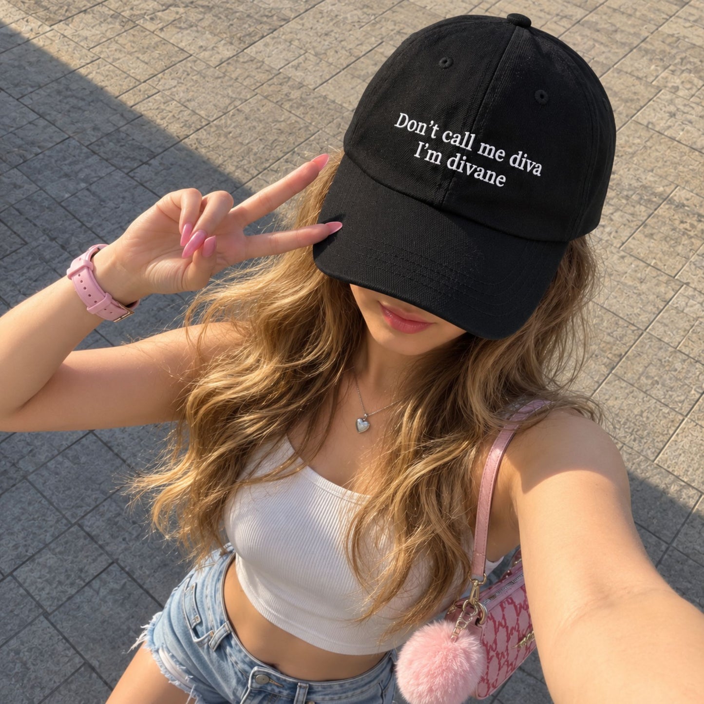 A woman outdoors wearing a black baseball cap with white embroidered text that reads 'Don't call me diva I'm divane'. She is posing for a selfie, making a peace sign with her hand.