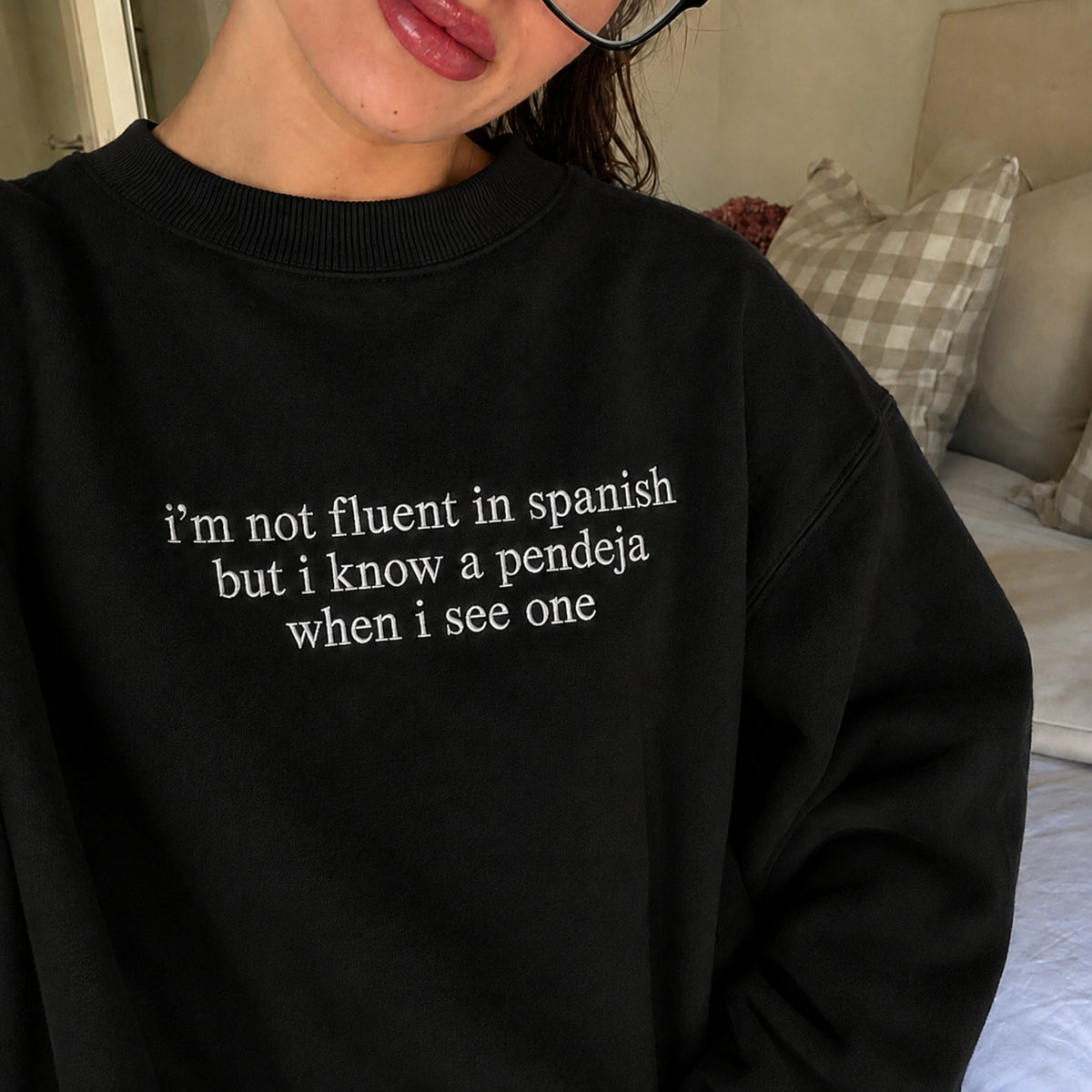 A person wearing a black crewneck sweatshirt with white text that reads 'i'm not fluent in spanish but i know a pendeja when i see one', photographed in a bedroom setting.