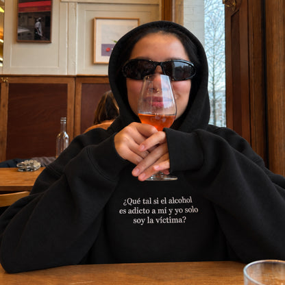 Person wearing a black hoodie and dark sunglasses holds a glass of wine at a restaurant, with the embroidered Spanish quote “¿Qué tal si el alcohol es adicto a mí y yo solo soy la víctima?” on the chest.