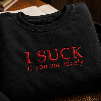A folded black crewneck sweatshirt with red embroidered text reading 'I SUCK if you ask nicely' on the front, shown on a warm brown textured surface with an open book in the background.