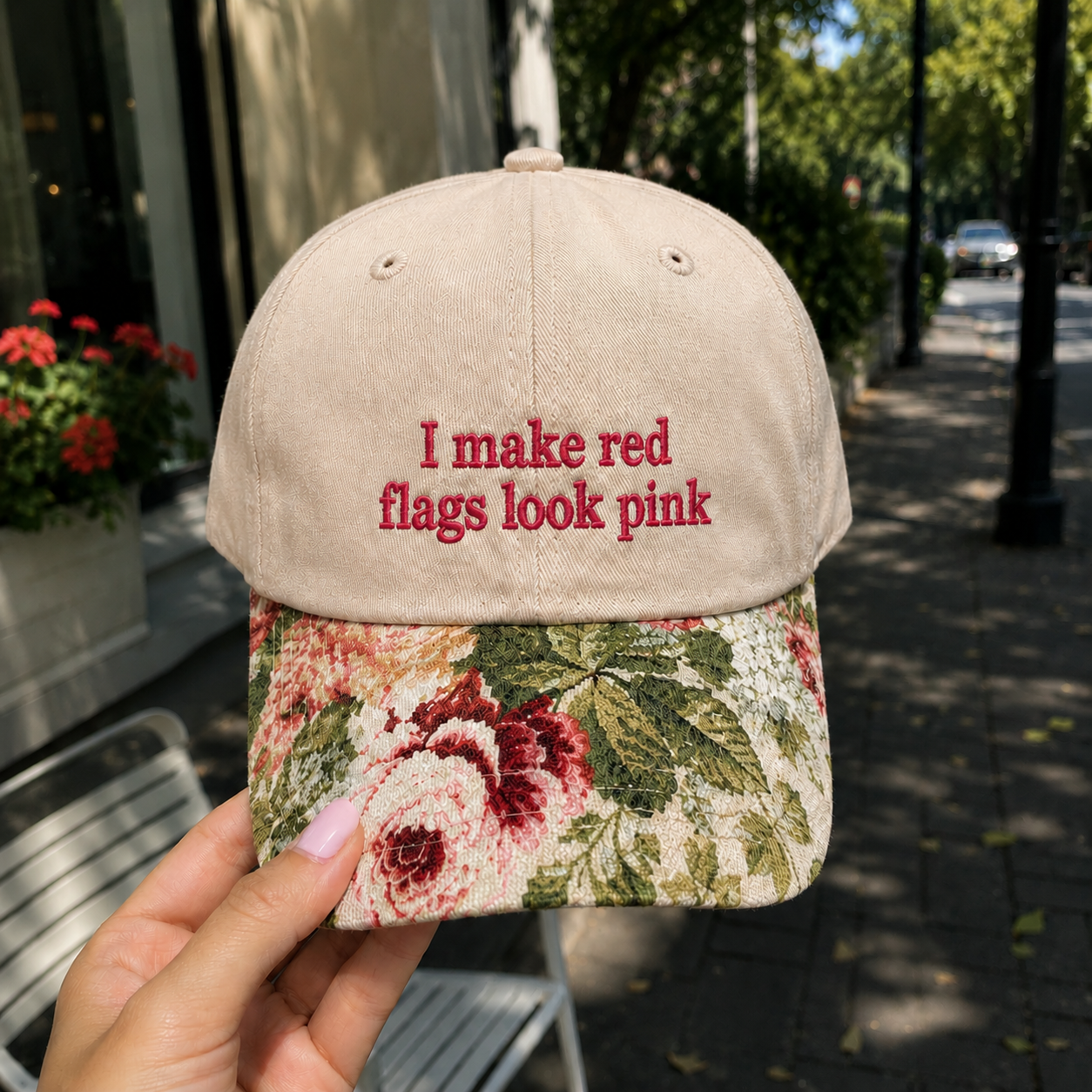 Close-up of a hand holding an embroidered 'I make red flags look pink' baseball hat with floral brim outdoors.
